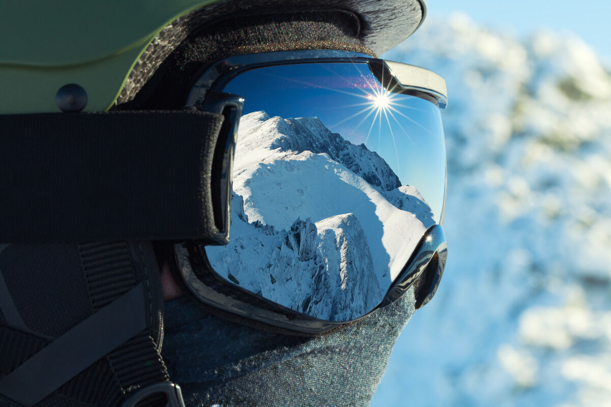 A side profile covered with a helmet, neck gaiter, and snow goggles. A snowcapped mountain is reflected in the goggles.